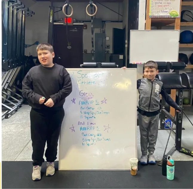 Two young Cynergy Kids athletes with the weekly spotlight athlete whiteboard